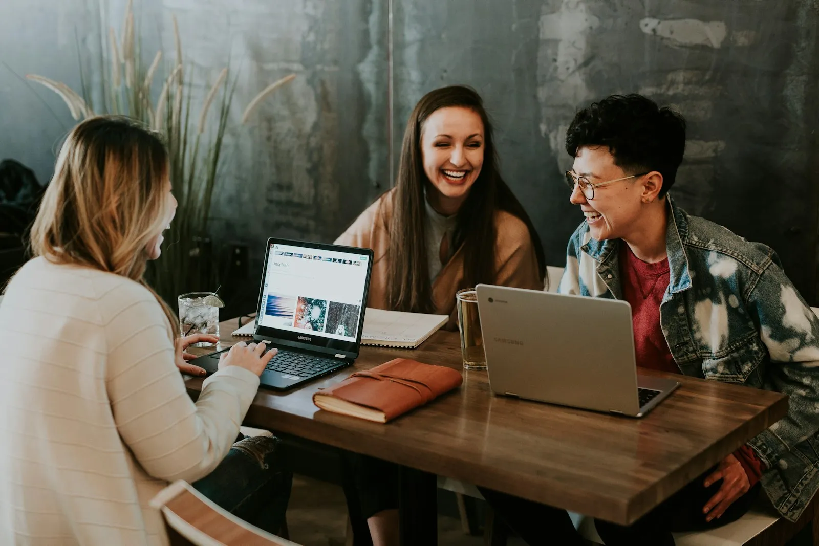 Three committee members laughing together over laptops and a leather notebook at a wooden table